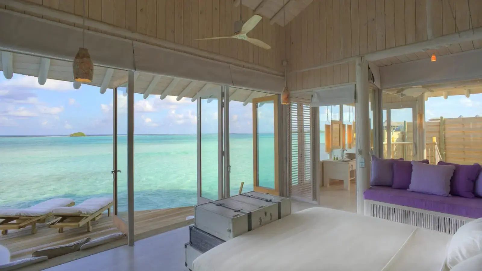 Helles Schlafzimmer einer Wasservilla mit Blick durch geöffnete Glastüren auf den Ozean.