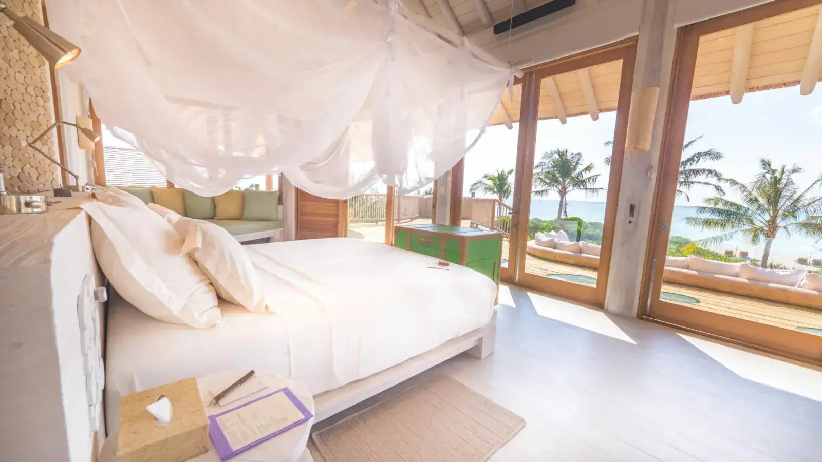 Helles Schlafzimmer mit Himmelsbett, grünem Schrank und Blick auf Palme und Meer.
