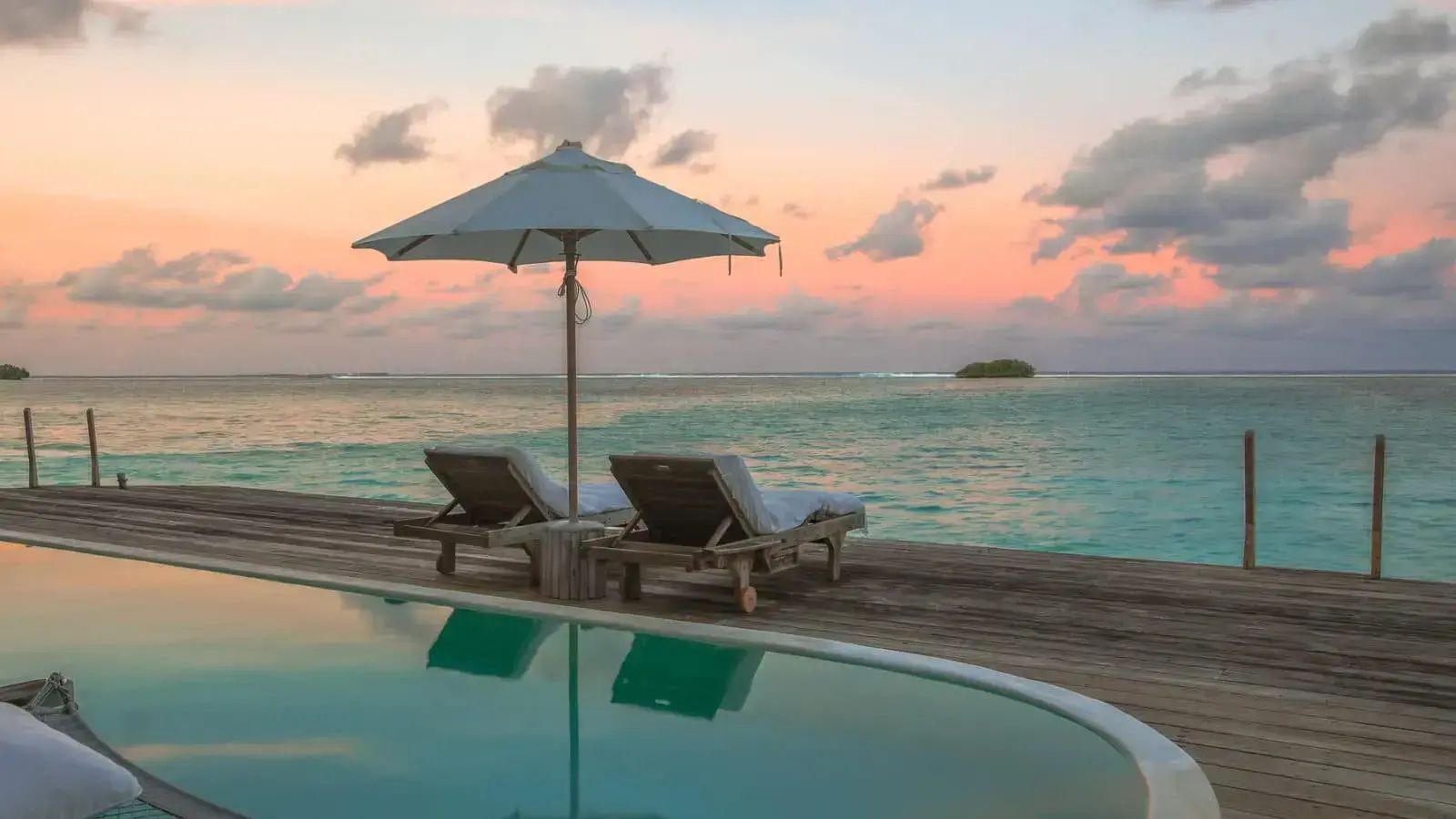 Sonnendeck am Pool mit zwei Liegestühlen und einem Sonnenschirm vor dem Meer bei Abendstimmung.