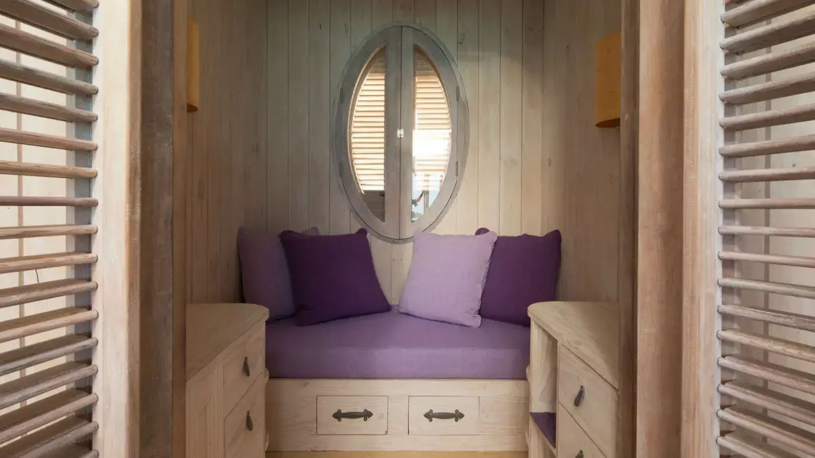In die Wand eingelassene Sitznische aus hellem Holz mit lilafarbenen Kissen und ovalem Fenster.