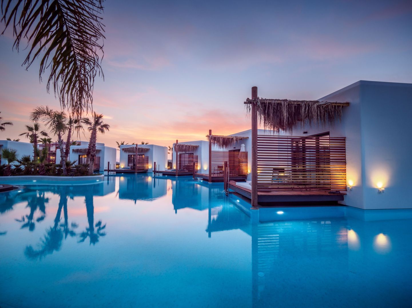 Bungalows mit Holzterrassen an einer großen Poollandschaft während der Abenddämmerung.