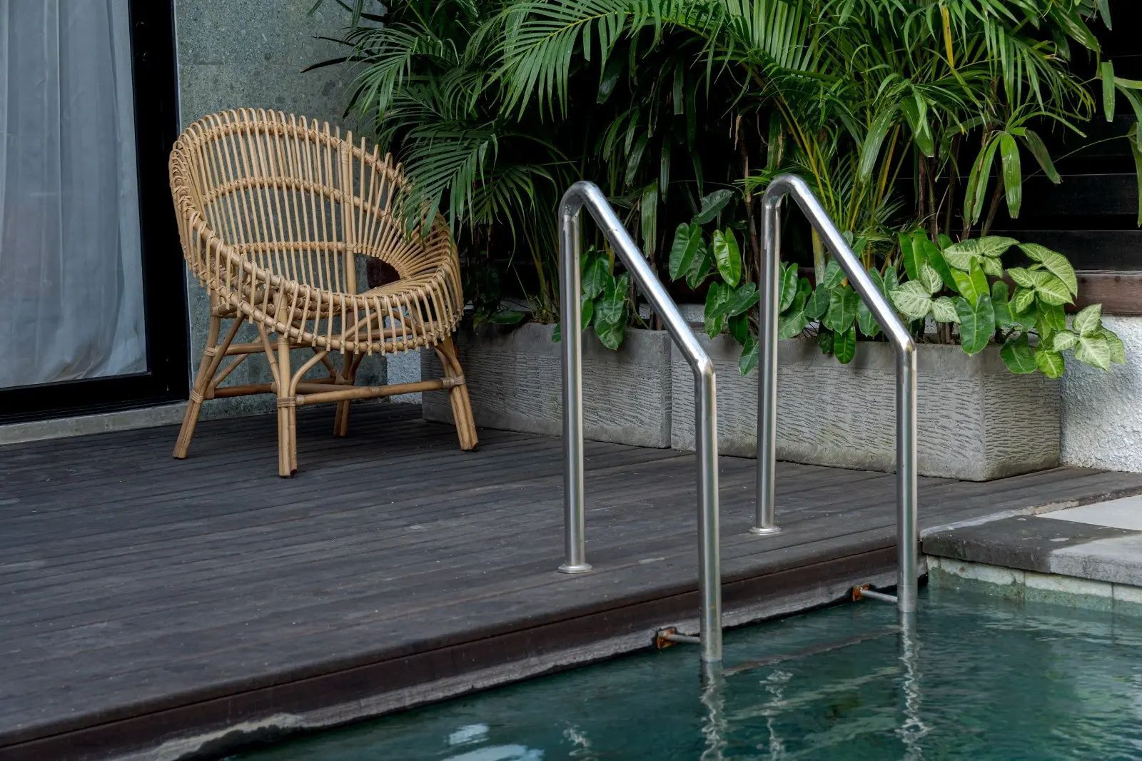 Edelstahlleiter am Poolrand neben einer Holzterrasse und einem Rattanstuhl.