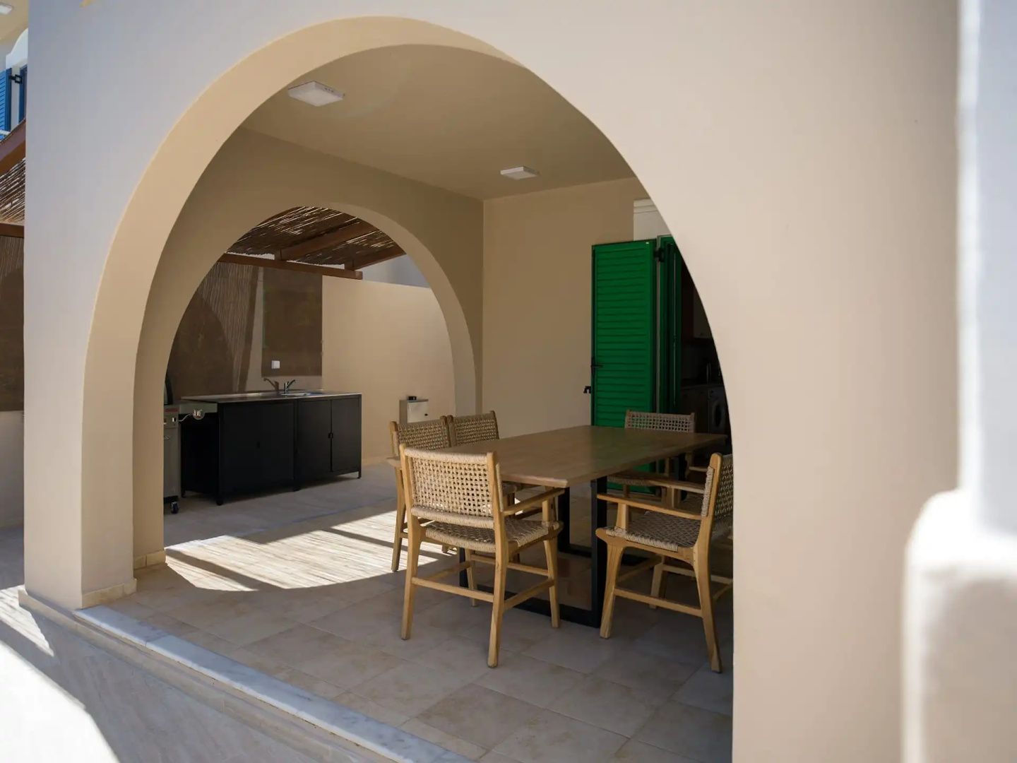 Überdachter Terrassenbereich mit Holztisch, Flechtstühlen und einer Außenküche im Hintergrund.