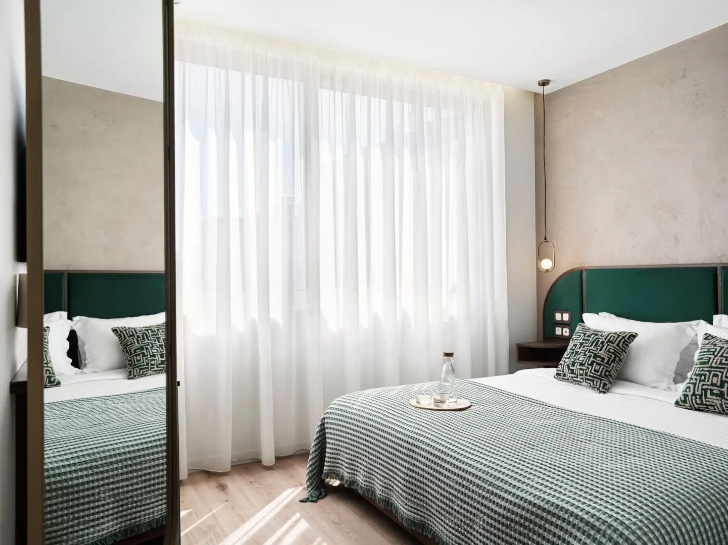 Helles Hotelzimmer mit Doppelbett, grünen Kopfteilen und großen transparenten Vorhängen am Fenster.