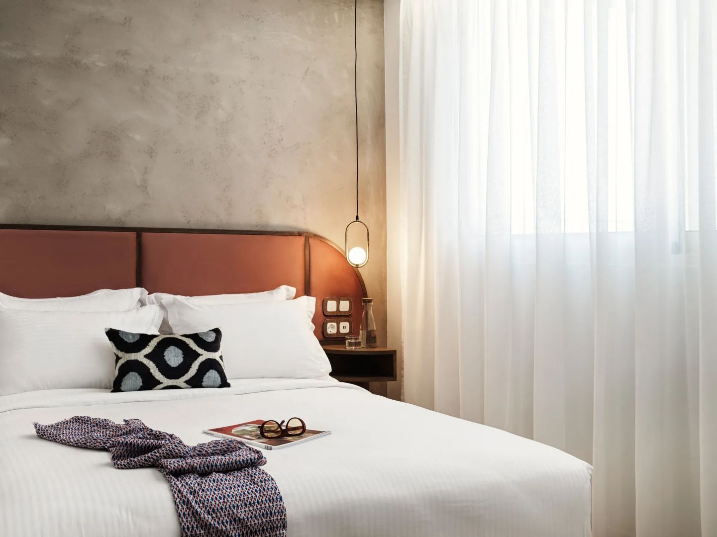 Doppelbett mit weißer Bettwäsche, rotbraunem Kopfteil und hängender Nachttischlampe am Fenster.