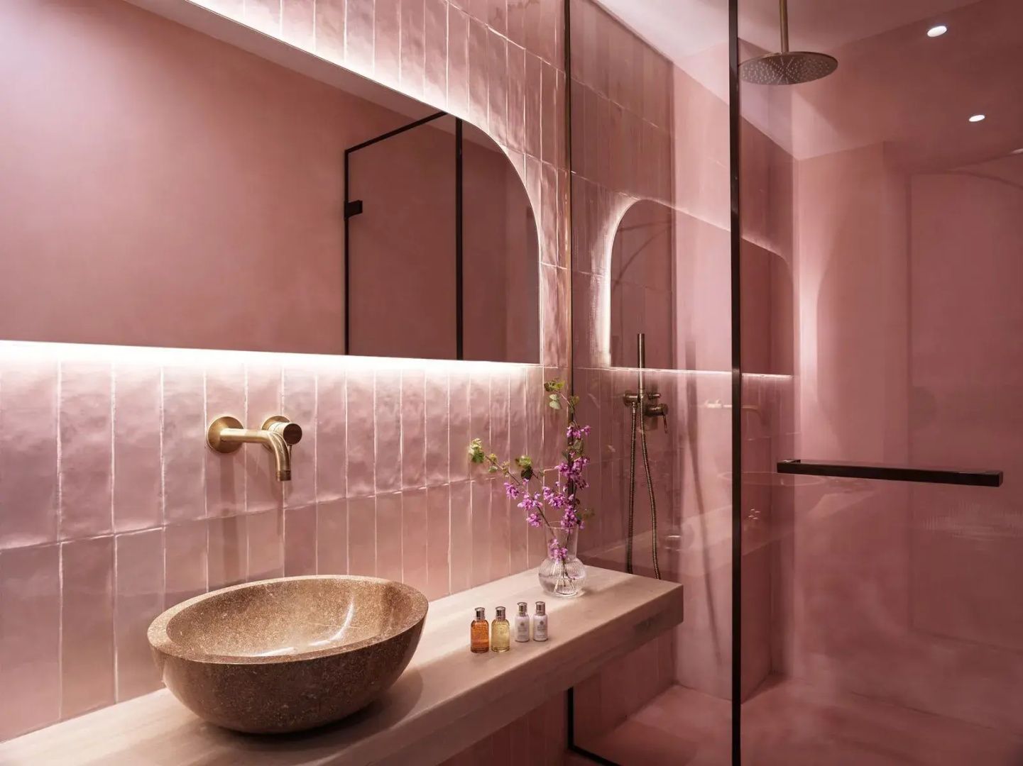 Badezimmer mit rosa Wandfliesen, beleuchtetem Spiegel, Waschtisch und Dusche hinter Glas.