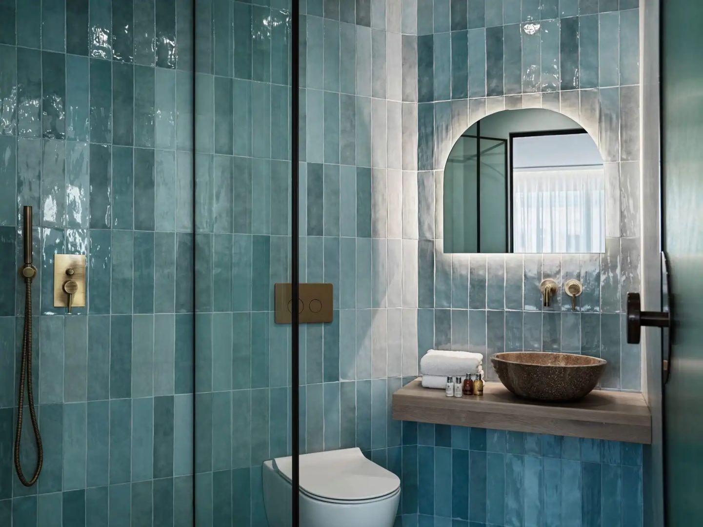 Badezimmer mit türkisblau gefliesten Wänden, rundem beleuchtetem Spiegel und Waschbecken.