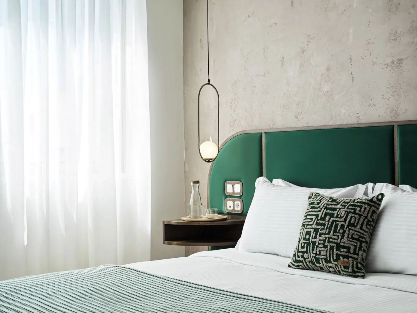Doppelbett mit grünem Kopfteil, weißer Bettwäsche und hängender Designerlampe.