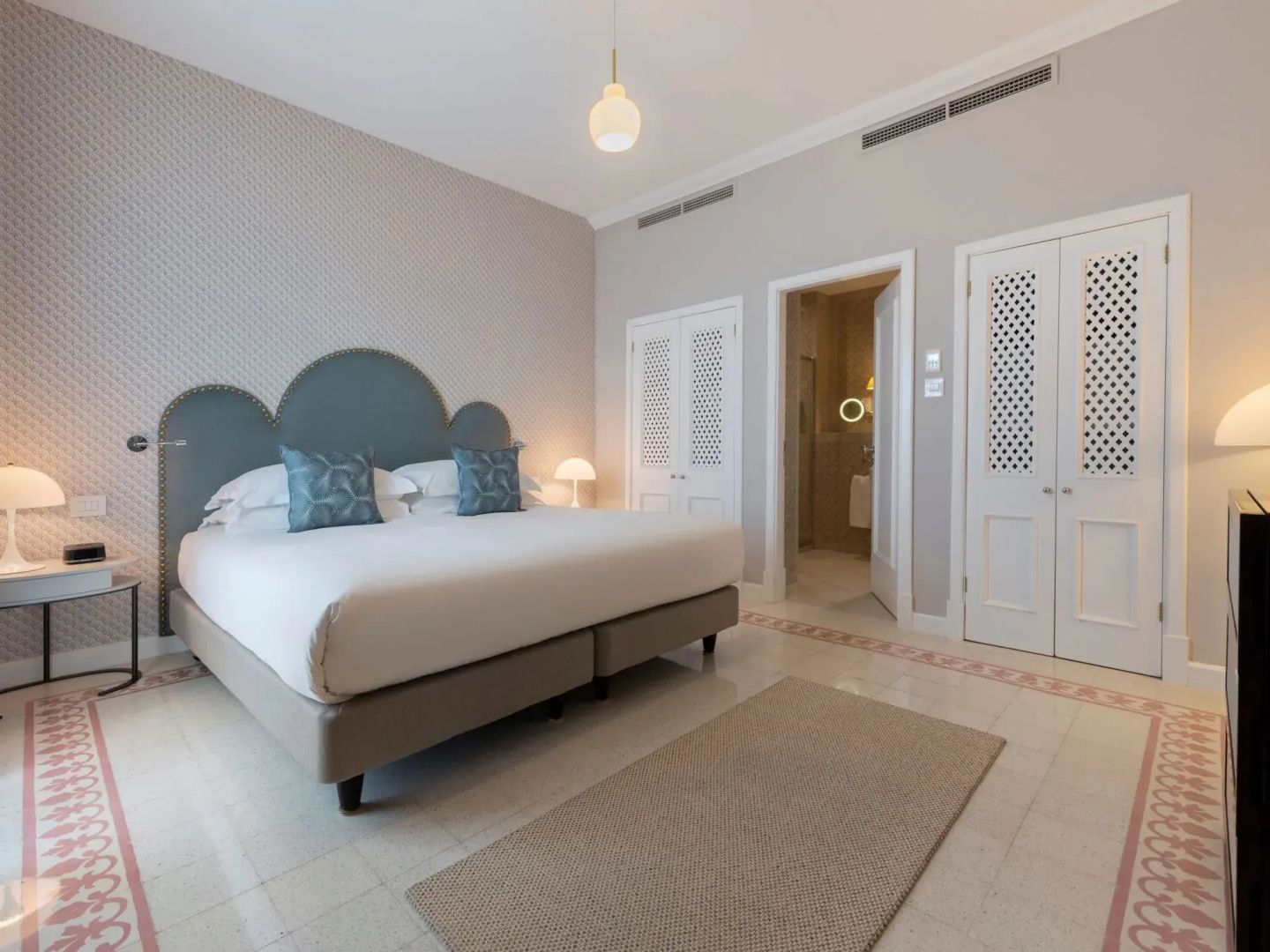 Hotelzimmer mit großem Doppelbett, blauem Kopfteil, hellen Fliesen und Durchgang zum Badezimmer.