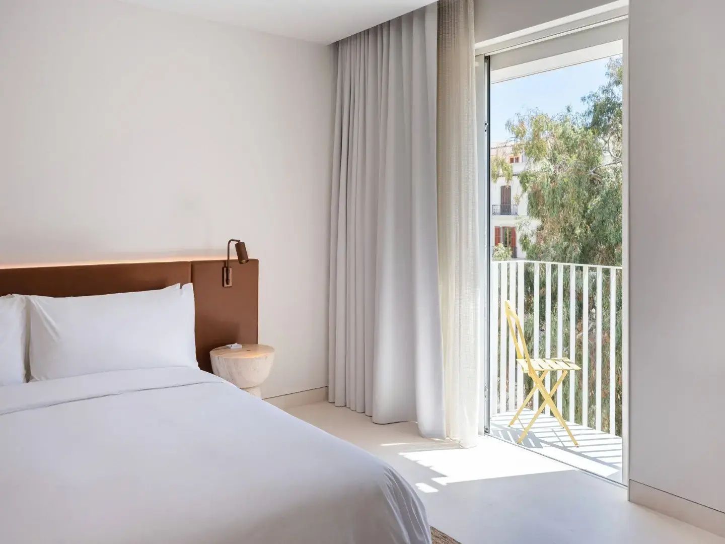 Helles Hotelzimmer mit weißem Bett, Nachttisch und Balkon mit gelbem Stuhl.