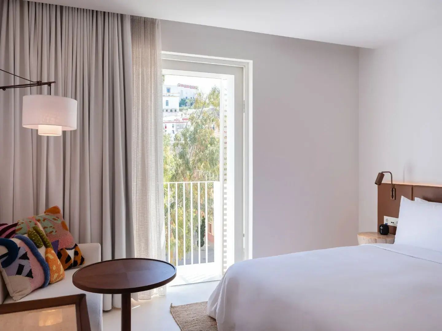 Helles Hotelzimmer mit weißem Bett, kleinem Holztisch und Blick durch die Balkontür auf Bäume.