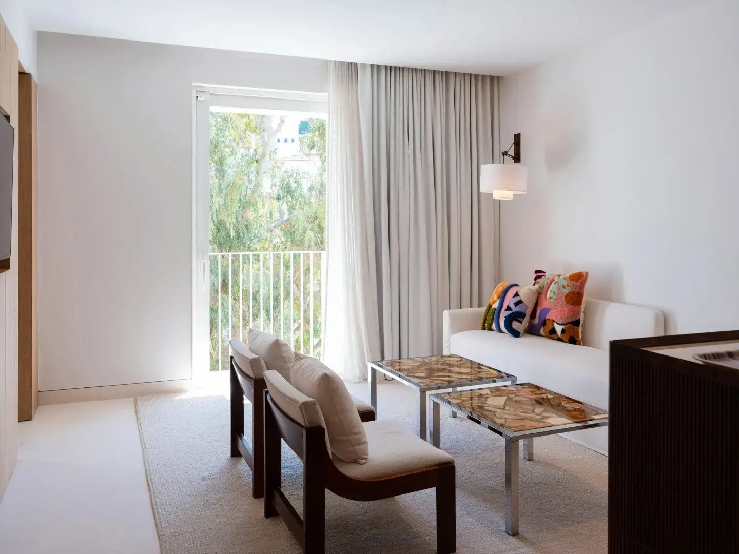 Helles Sitzzimmer mit weißem Sofa, bunten Kissen, zwei Holzsesseln und großem Fenster zum Balkon.