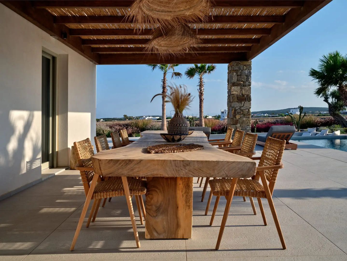 Überdachte Terrasse mit langem Holztisch, Stühlen und Blick auf Palmen.