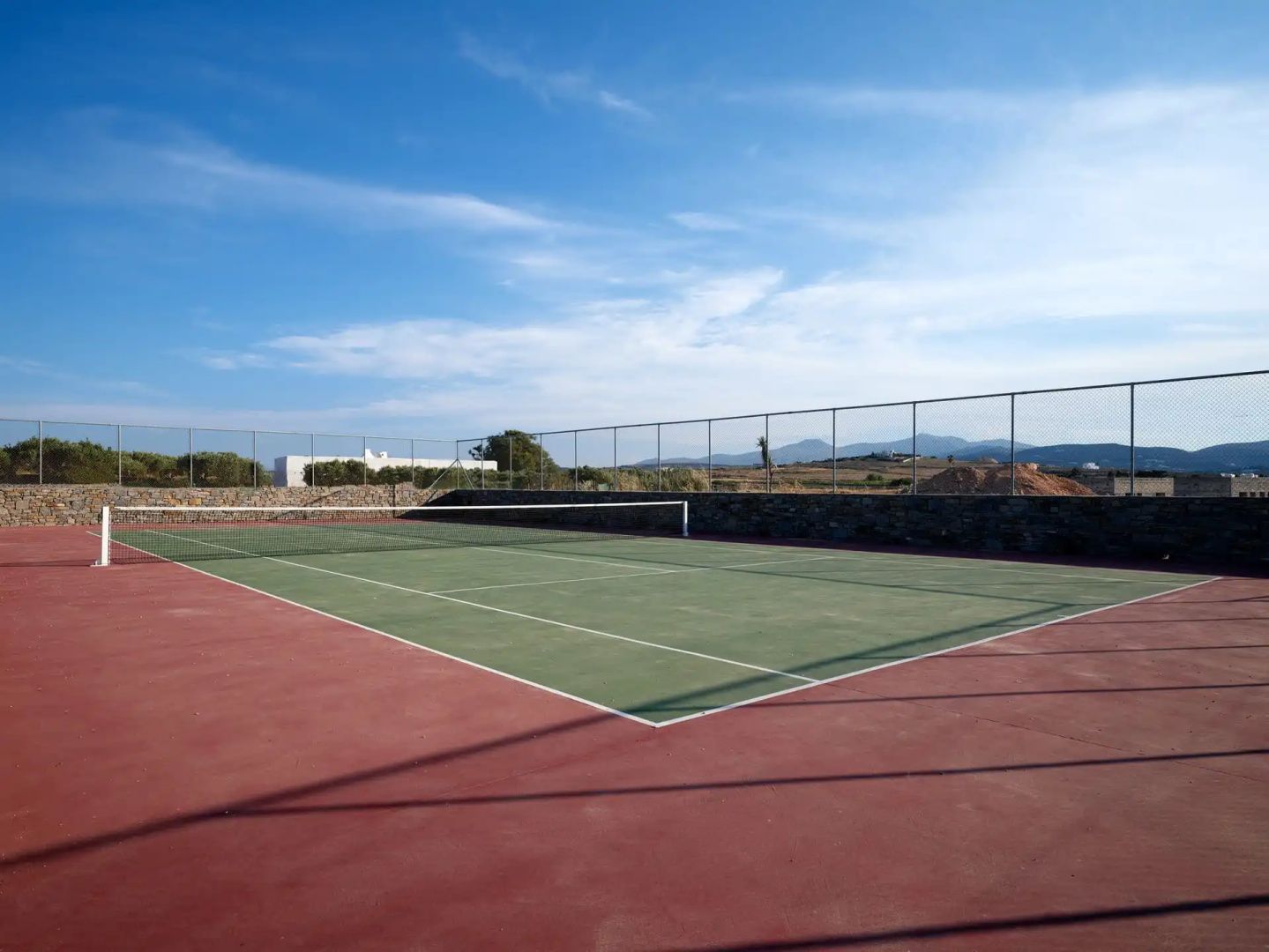 Tennisplatz im Freien mit roter Umrandung und grüner Spielfläche vor blauem Himmel.