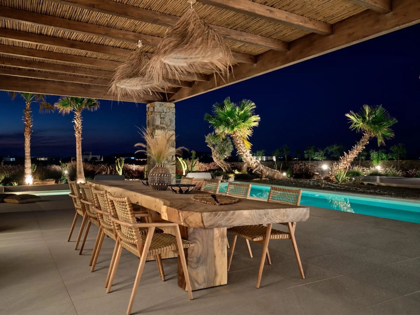 Überdachter Esstisch aus Holz mit Stühlen auf der Terrasse neben dem beleuchteten Pool.