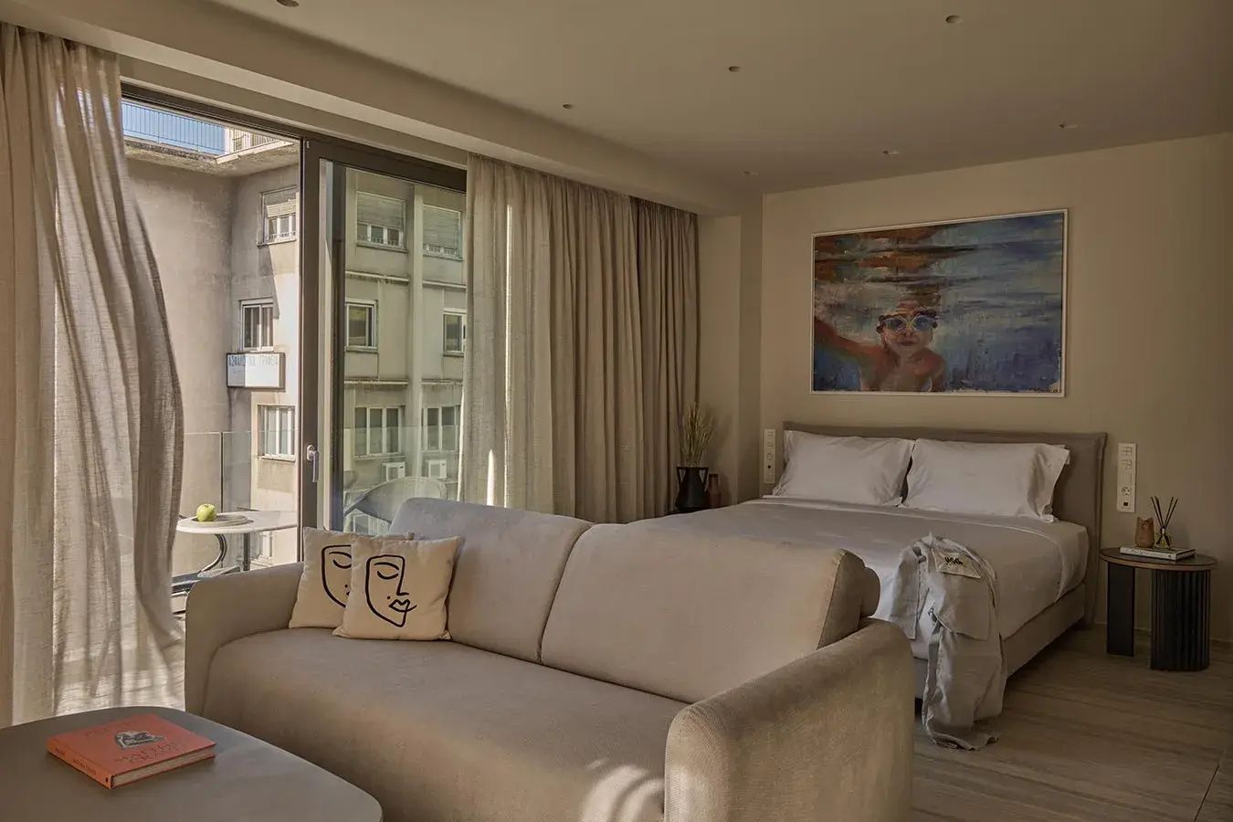 Modernes Hotelzimmer mit hellgrauem Sofa, Doppelbett und Fensterfront zum Stadtbereich.