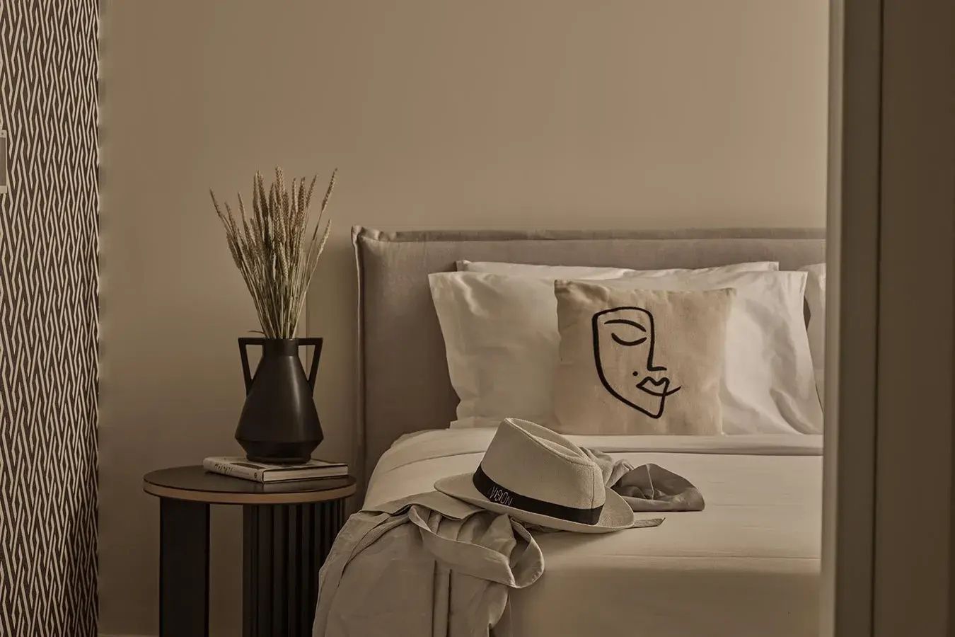 Kissen mit Gesichtszeichnung auf dem Bett neben Nachttisch mit dunkler Vase und Trockenblumen.