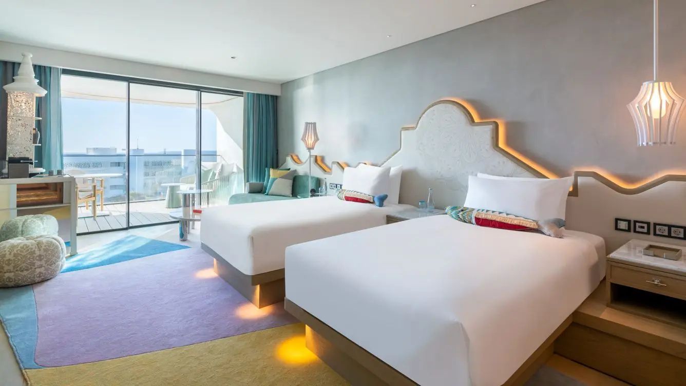 Modernes Hotelzimmer mit zwei Einzelbetten, bunter Beleuchtung am Kopfteil und Balkonzugang.