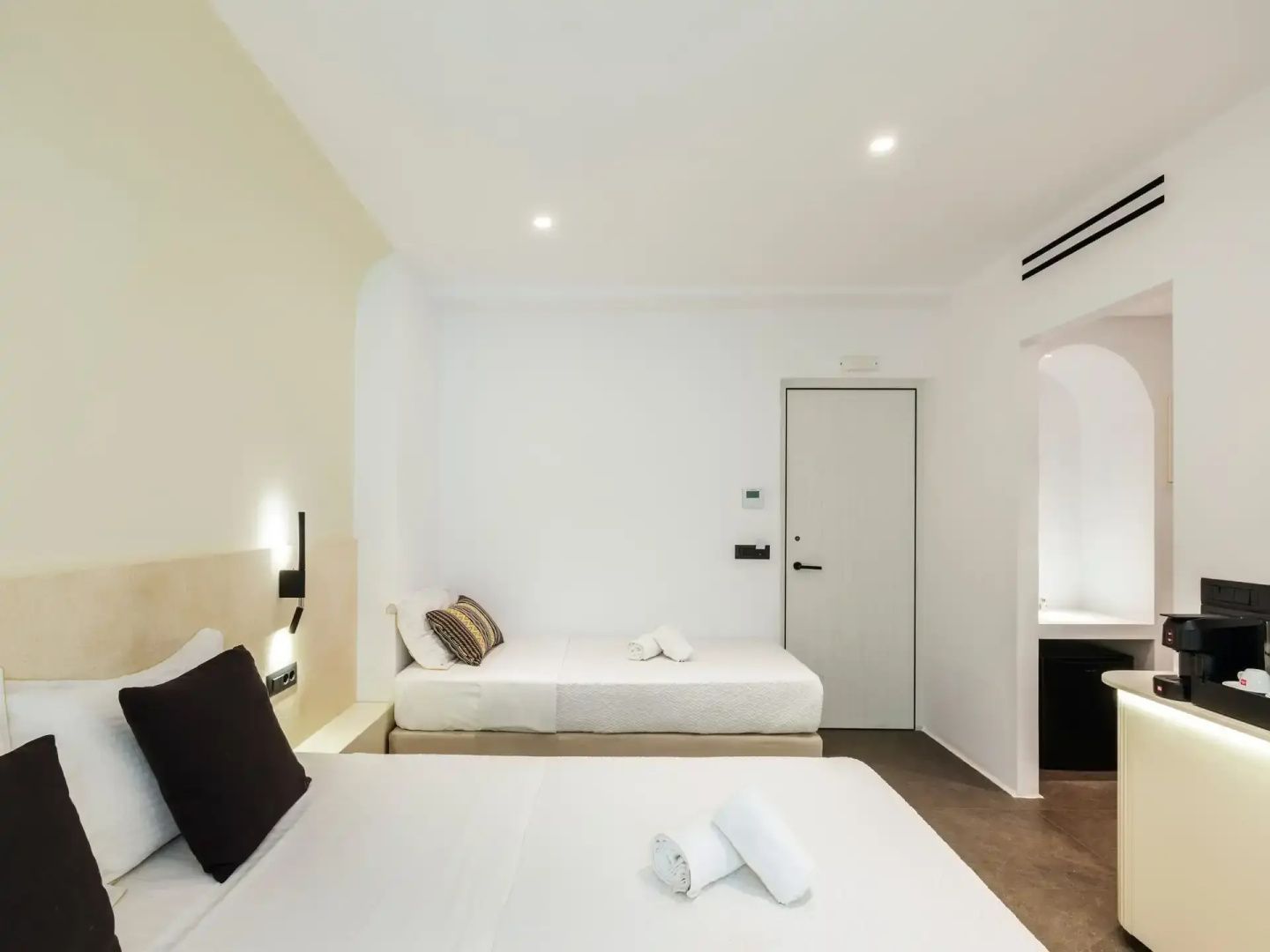Minimalistisch eingerichtetes Hotelzimmer mit Doppelbett und Einzelbett an weißer Wand.