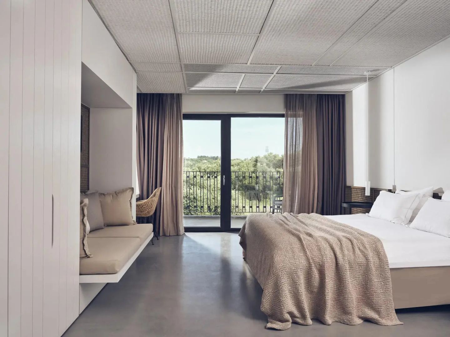 Helles minimalistisches Hotelzimmer mit Doppelbett, Sitzbank und Balkontür mit Blick ins Grüne.