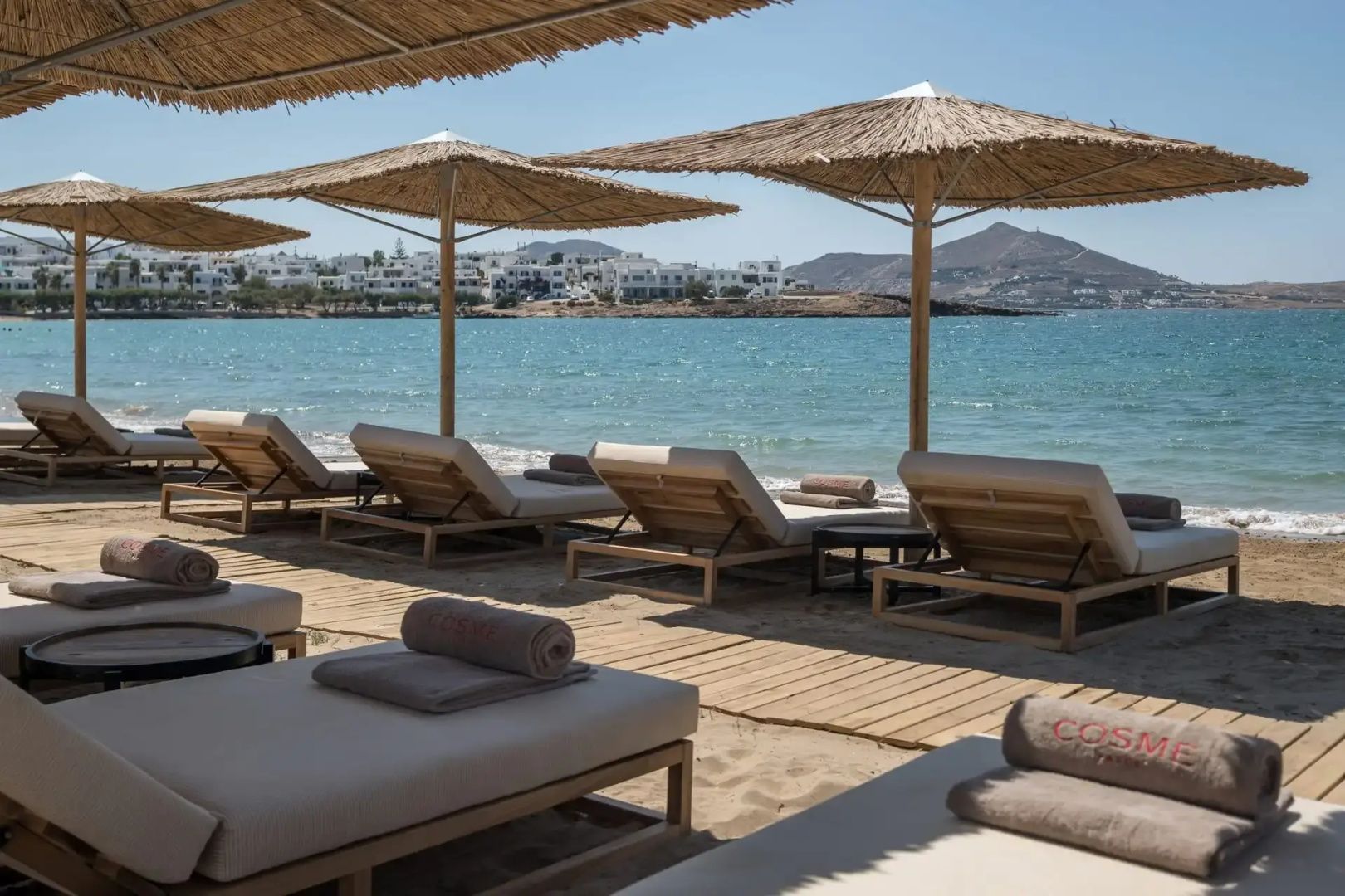 Querformat Alt-Text: Strandliegen unter Schilfschirmen am Sandstrand auf Paros mit türkisfarbener Bucht und Hügeln im Hintergrund