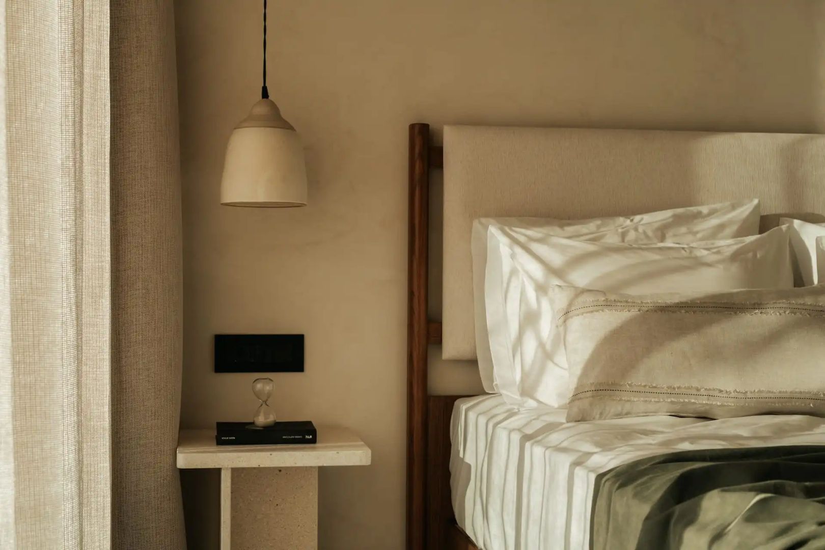 Bett mit beigefarbener Bettwäsche, Nachttisch aus Stein und hängender Lampe an der Wand.