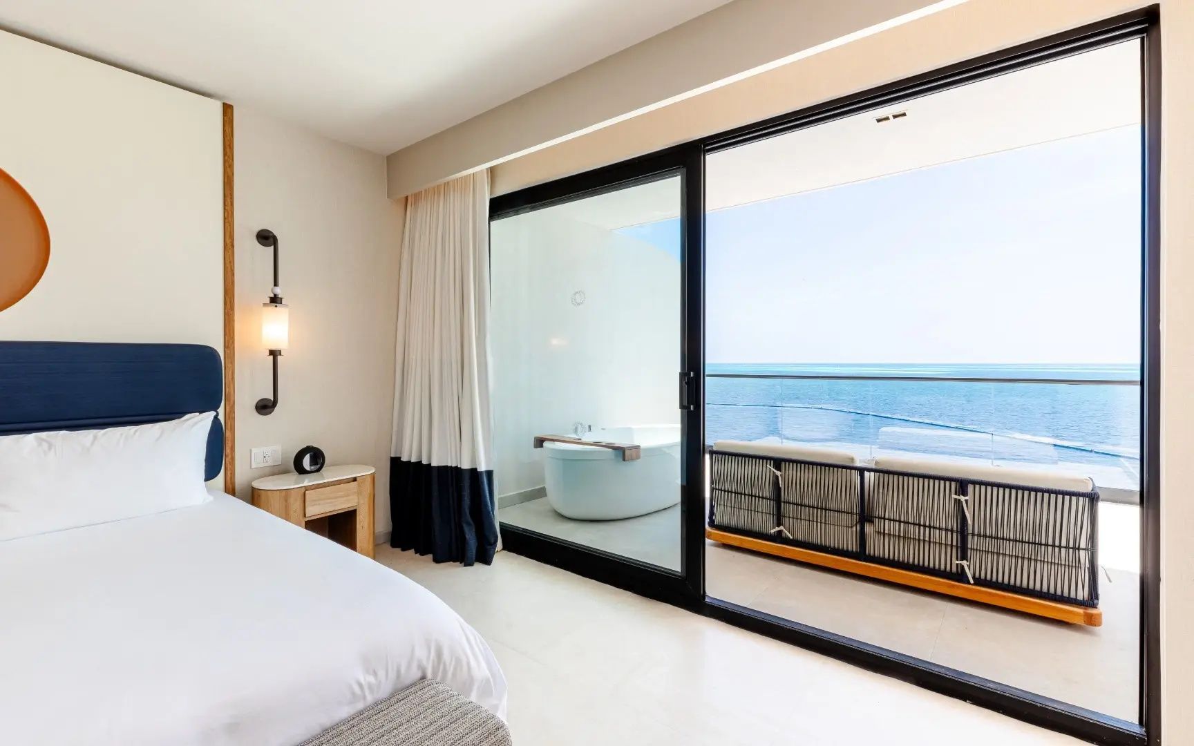 Blick durch eine Glasschiebetür auf einen Balkon mit Badewanne und Meerblick.