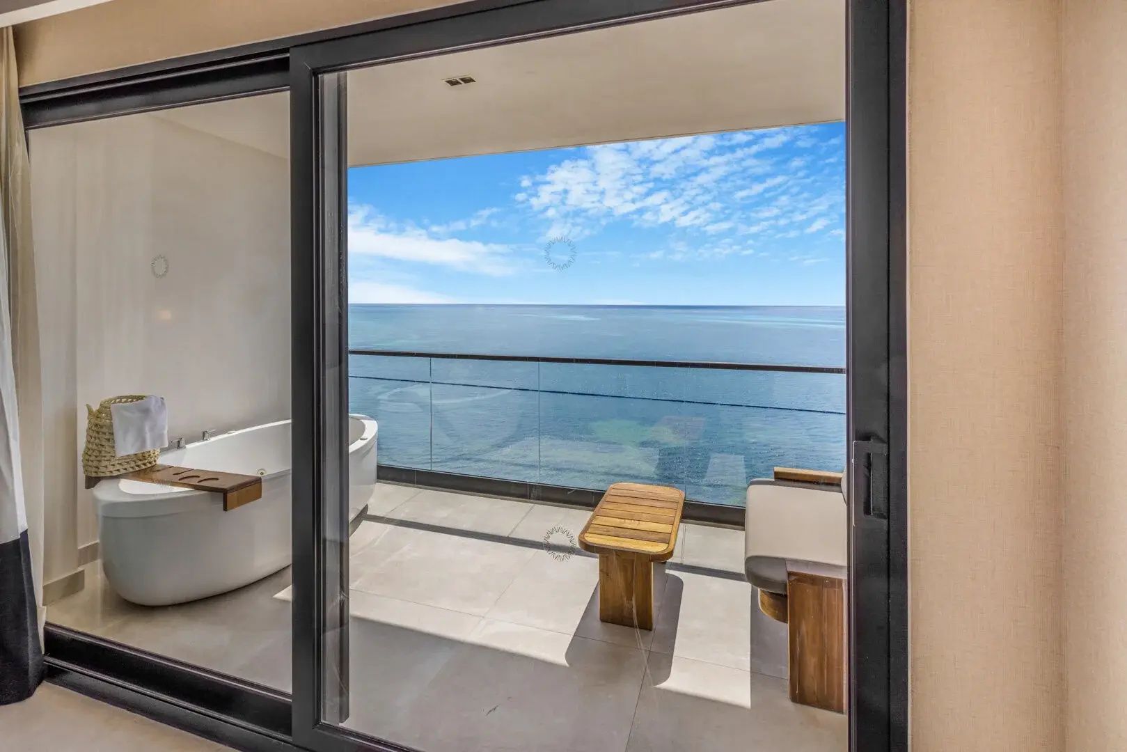 Blick durch die geöffnete Balkontür auf die Terrasse mit Badewanne und das blaue Meer.