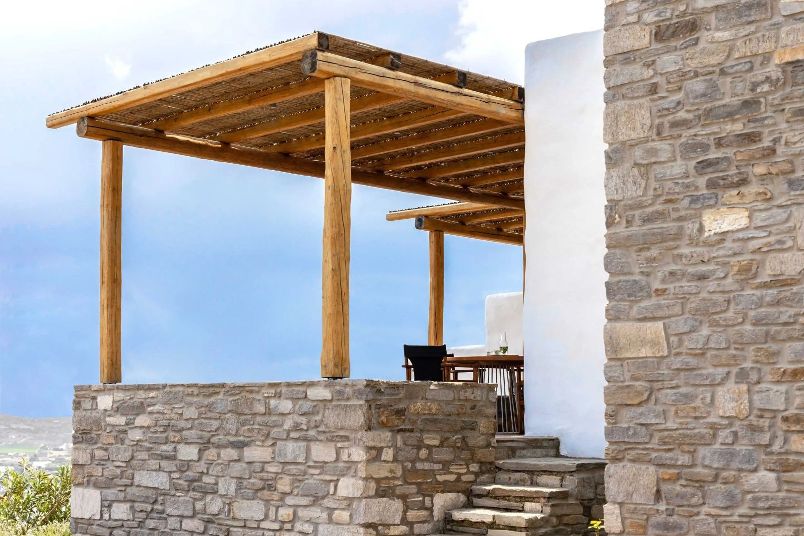 Außenbereich einer Villa mit Natursteinmauer, Holzpergola und Stufen unter blauem Himmel.