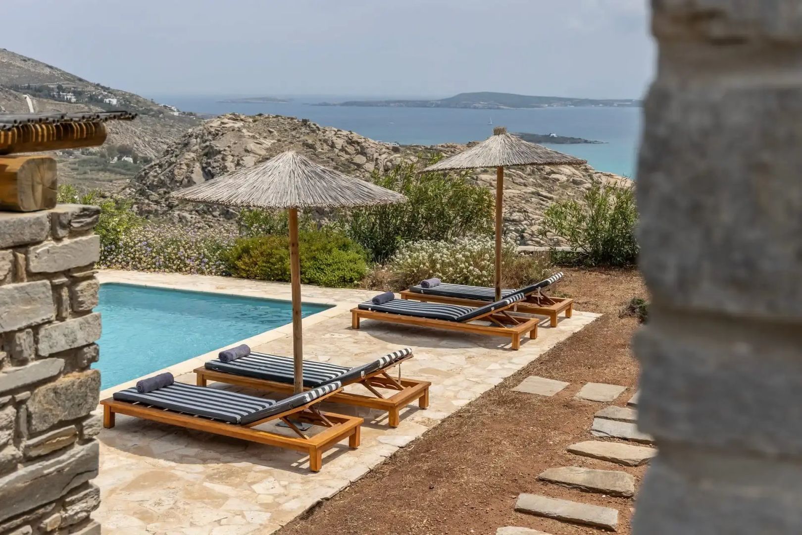 Sonnendeck am Pool mit Holzliegen, Reetdachschirmen und Blick auf das Meer und die Hügel.