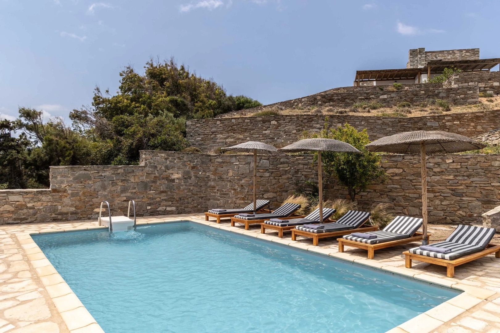 Poolanlage mit gepolsterten Liegestühlen und Sonnenschirmen vor einer Steinwand.
