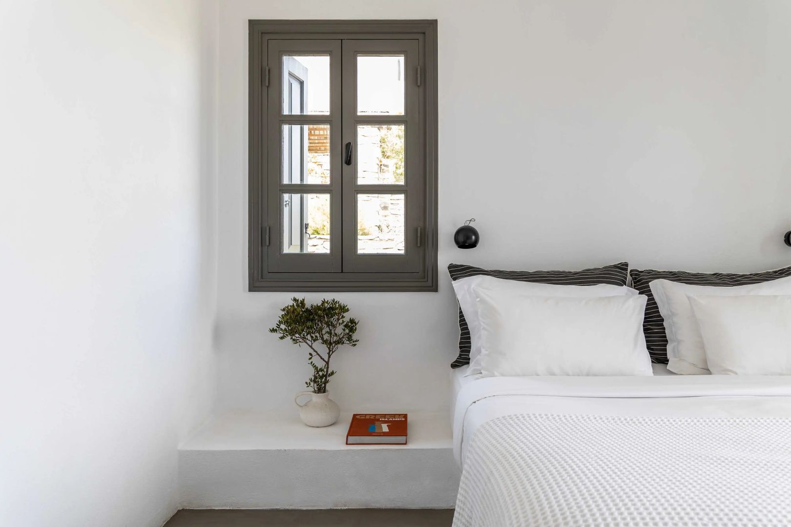 Weißes Bett neben einer Ablage mit Pflanze und kleinem Holzfenster an der Wand.