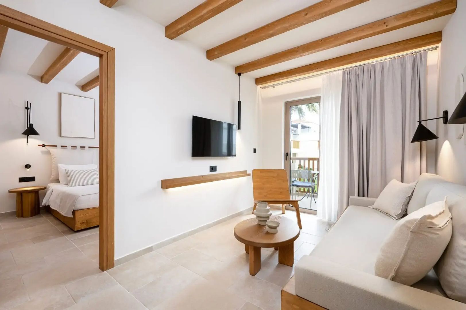 Hell eingerichtetes Wohnzimmer mit Holzdecke, Fernseher, Stuhl und Zugang zum Balkon.