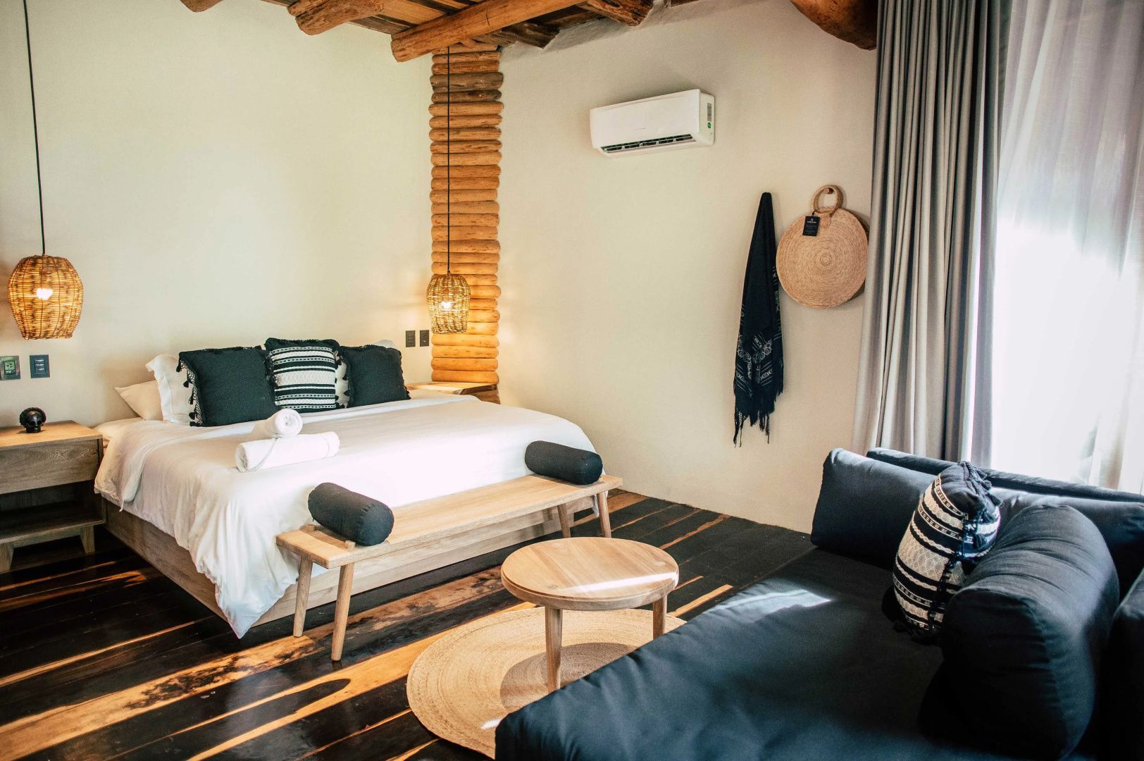 Helles Zimmer mit Doppelbett, hölzerner Sitzbank, Beistelltisch und dunkler Couch.