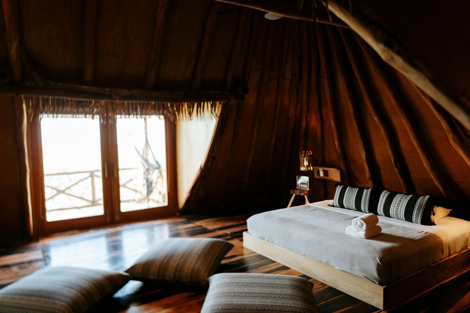 Zimmer unter einem Reetdach mit tiefem Bett, Sitzkissen und Holzboden.