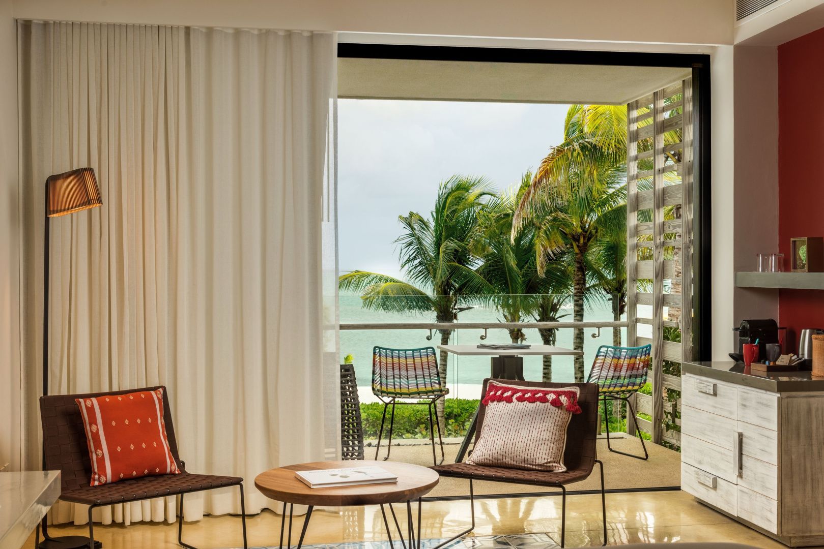 Blick aus dem Hotelzimmer auf den Balkon mit bunten Stühlen, Palmen und dem Meer.