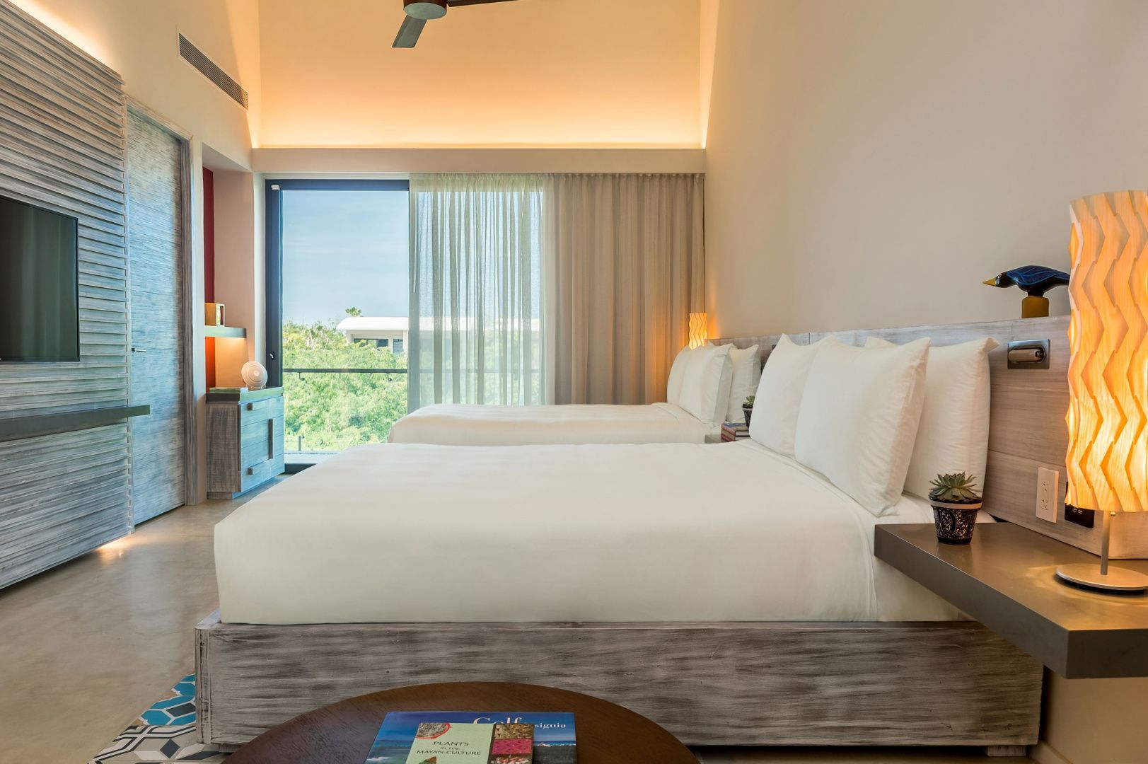 Zwei große Betten in einem hellen Hotelzimmer mit hoher Decke und Vorhängen am Fenster.