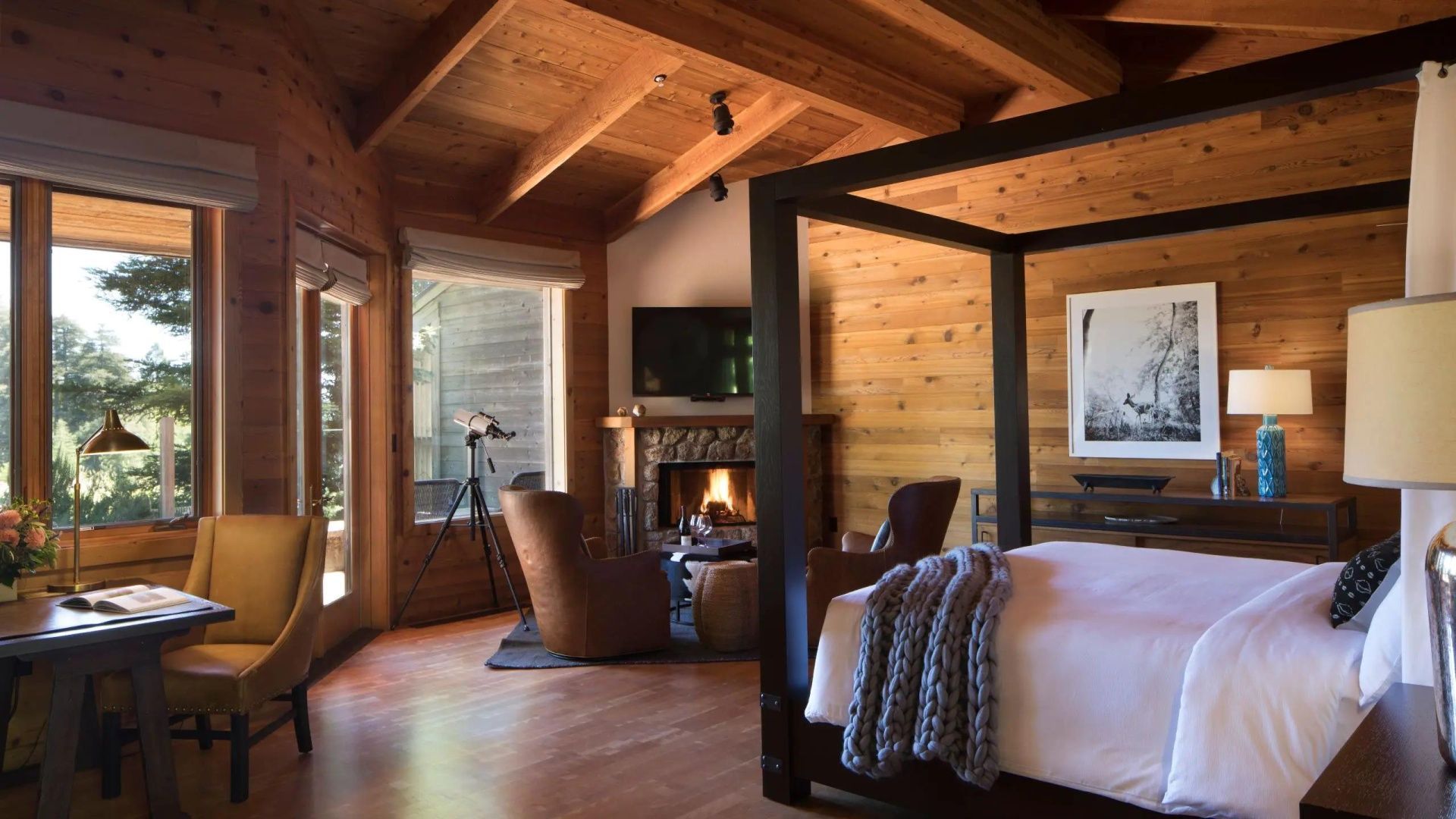 Holzverkleidetes Hotelzimmer mit Himmelbett, Kaminfeuer und Ledersesseln sowie Fernrohr am Fenster.