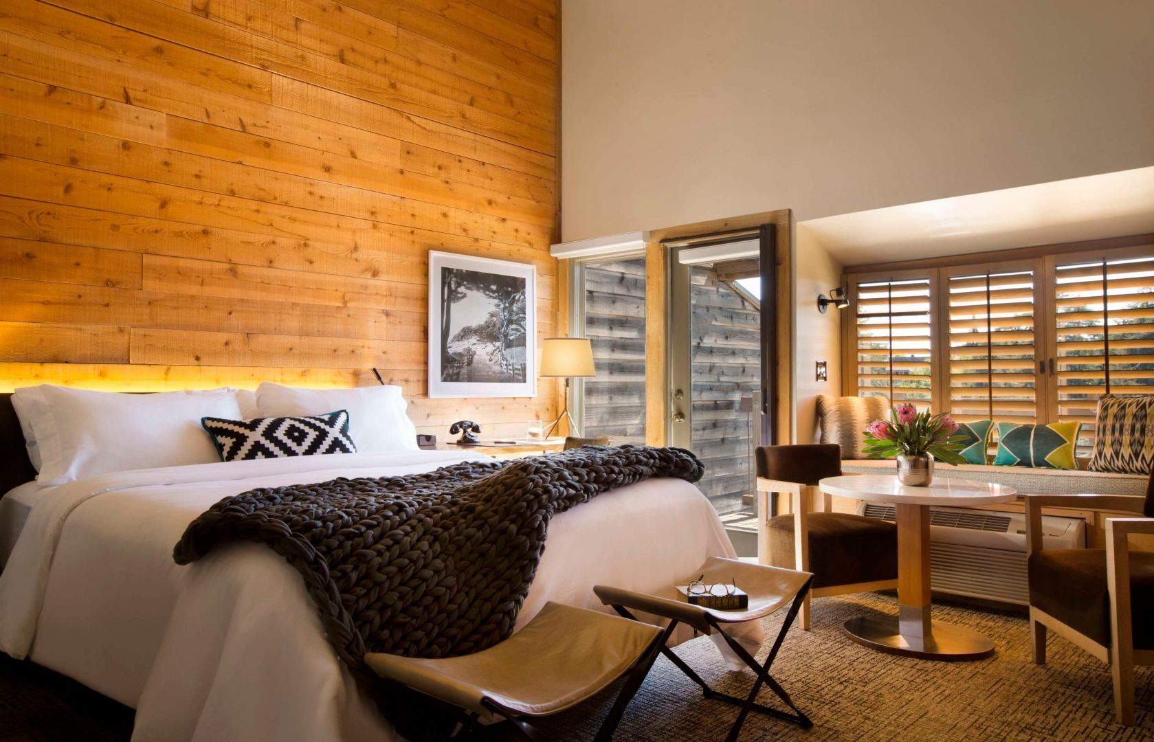 Schlafzimmer mit holzverkleideter Wand, Doppelbett, Sitzbereich und gläserner Balkontür.