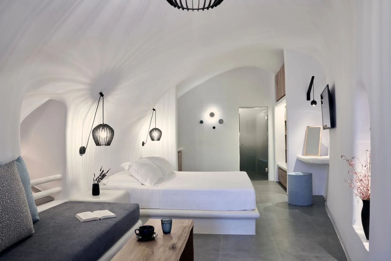 Weißes Höhlenzimmer mit Doppelbett, Gewölbedecke, modernen Wandleuchten und Sitzgelegenheit.