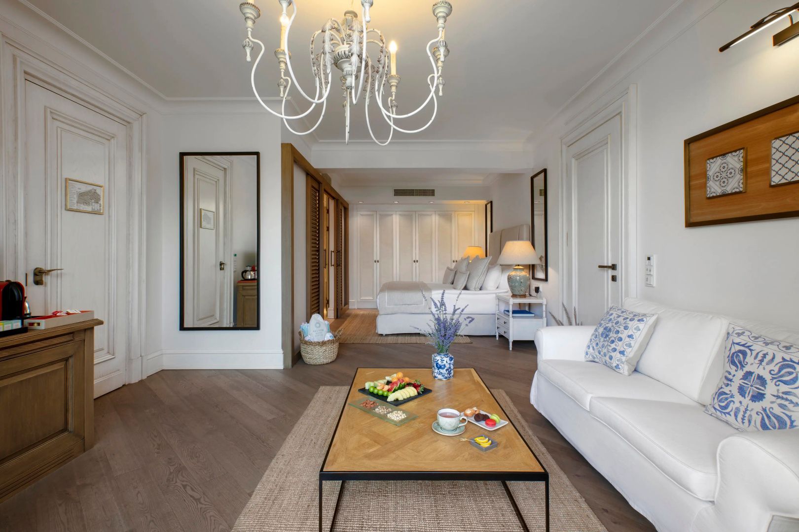 Wohnbereich einer Hotel-Suite mit Holztisch, Sofa, Kronleuchter und Bett im Hintergrund.
