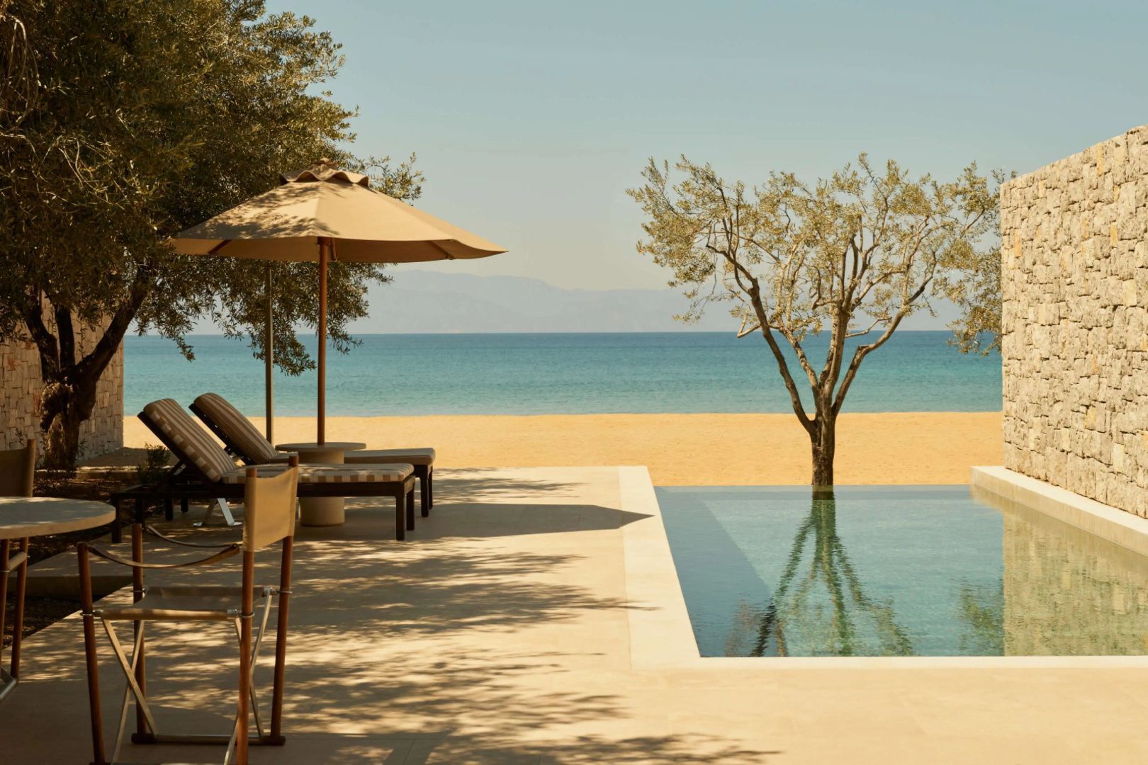 Sonnenschirm und Liegen neben einem Pool direkt am Sandstrand und blauen Meer.
