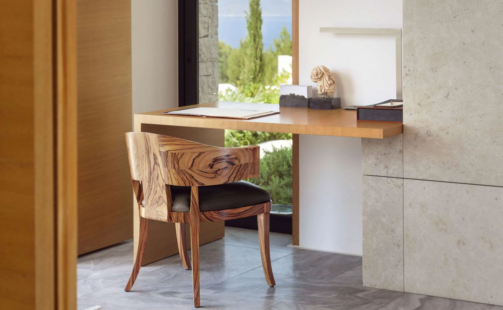 Schreibtisch aus Holz mit einem gemusterten Stuhl und Blick durch das Fenster ins Grüne.