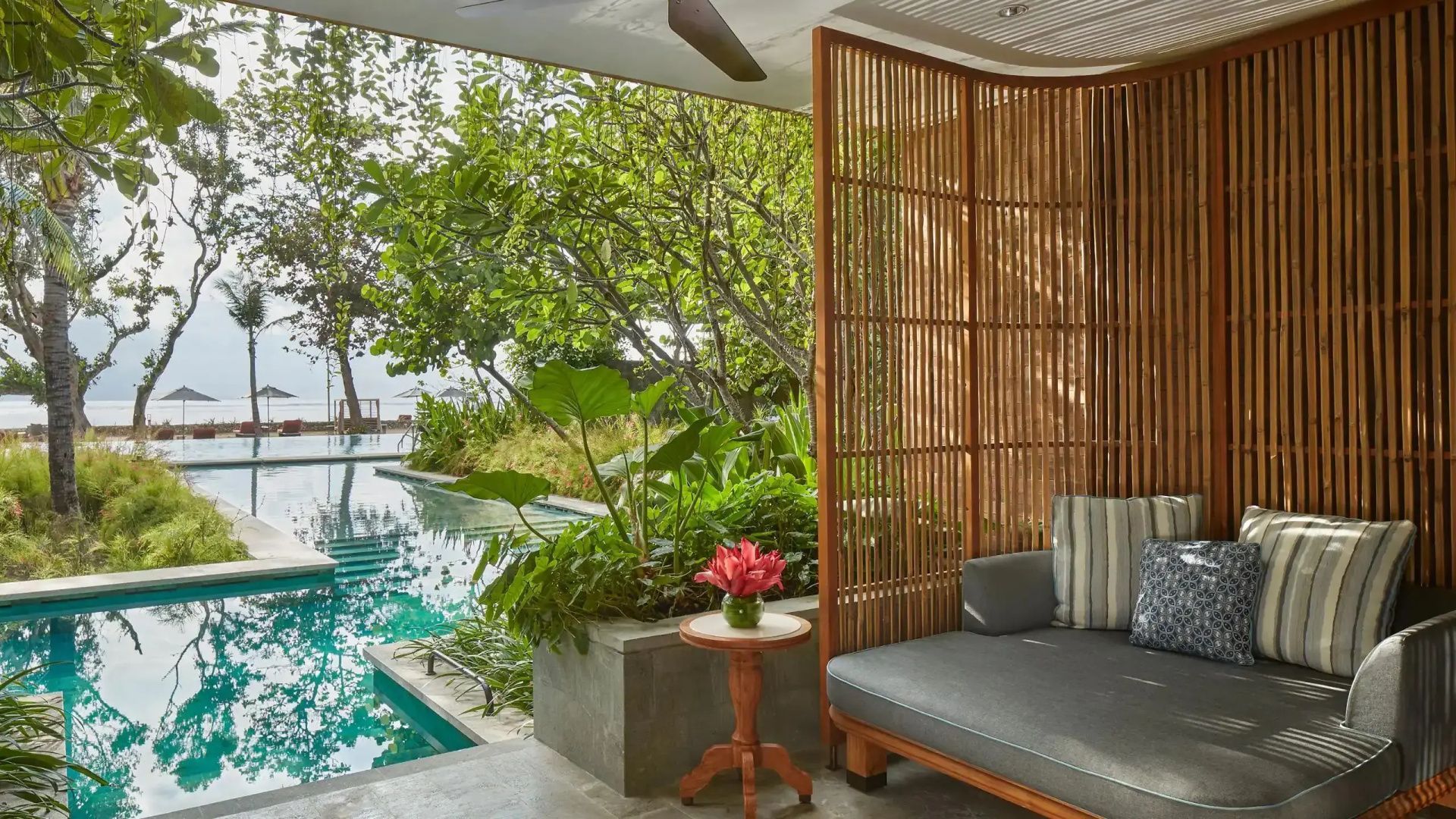 Überdachte Terrasse mit Holzparavent, Tagesbett und Ausblick auf Grünpflanzen und einen Pool.