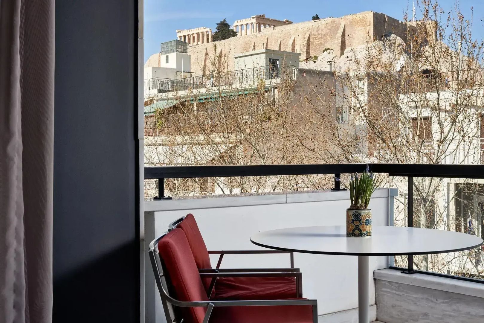 Balkon mit Tisch und roten Stühlen sowie Blick auf die Akropolis im Hintergrund.