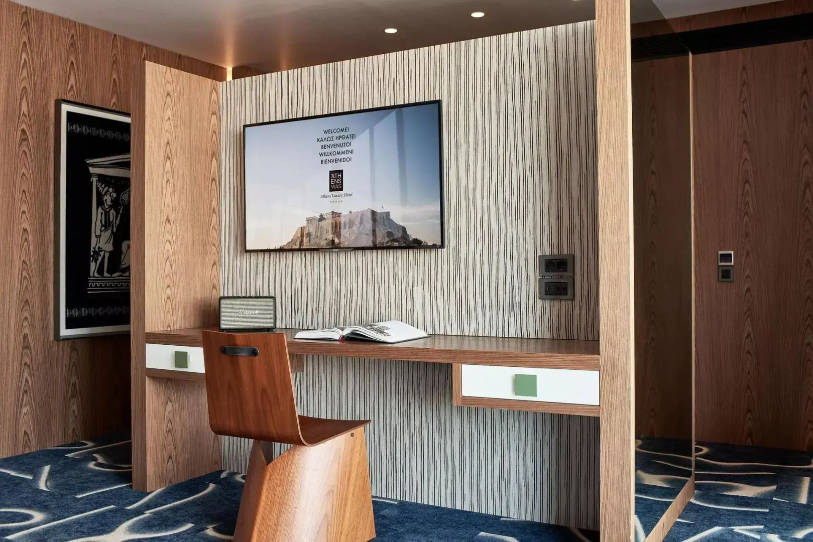 Holzschreibtisch mit Stuhl und einem Wandfernseher vor einer hellen Musterwand.