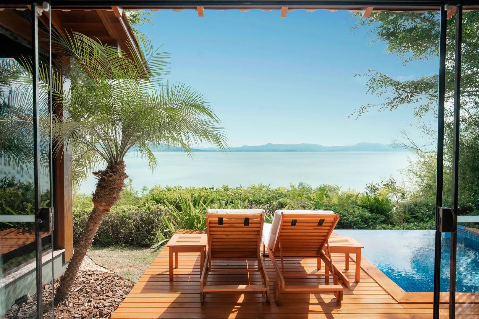 Zwei Holzliegen auf einer Holzplattform mit weitem Blick über das ruhige Meer und Berge am Horizont.