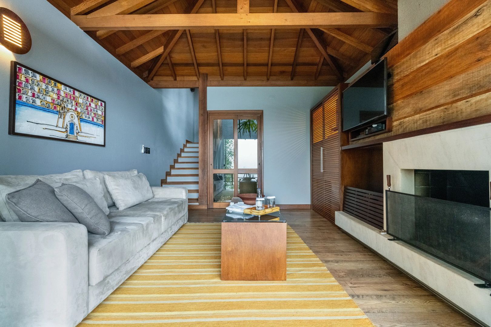 Wohnbereich mit grauem Sofa, gelbem Teppich, Fernseher und hoher Holzdecke.