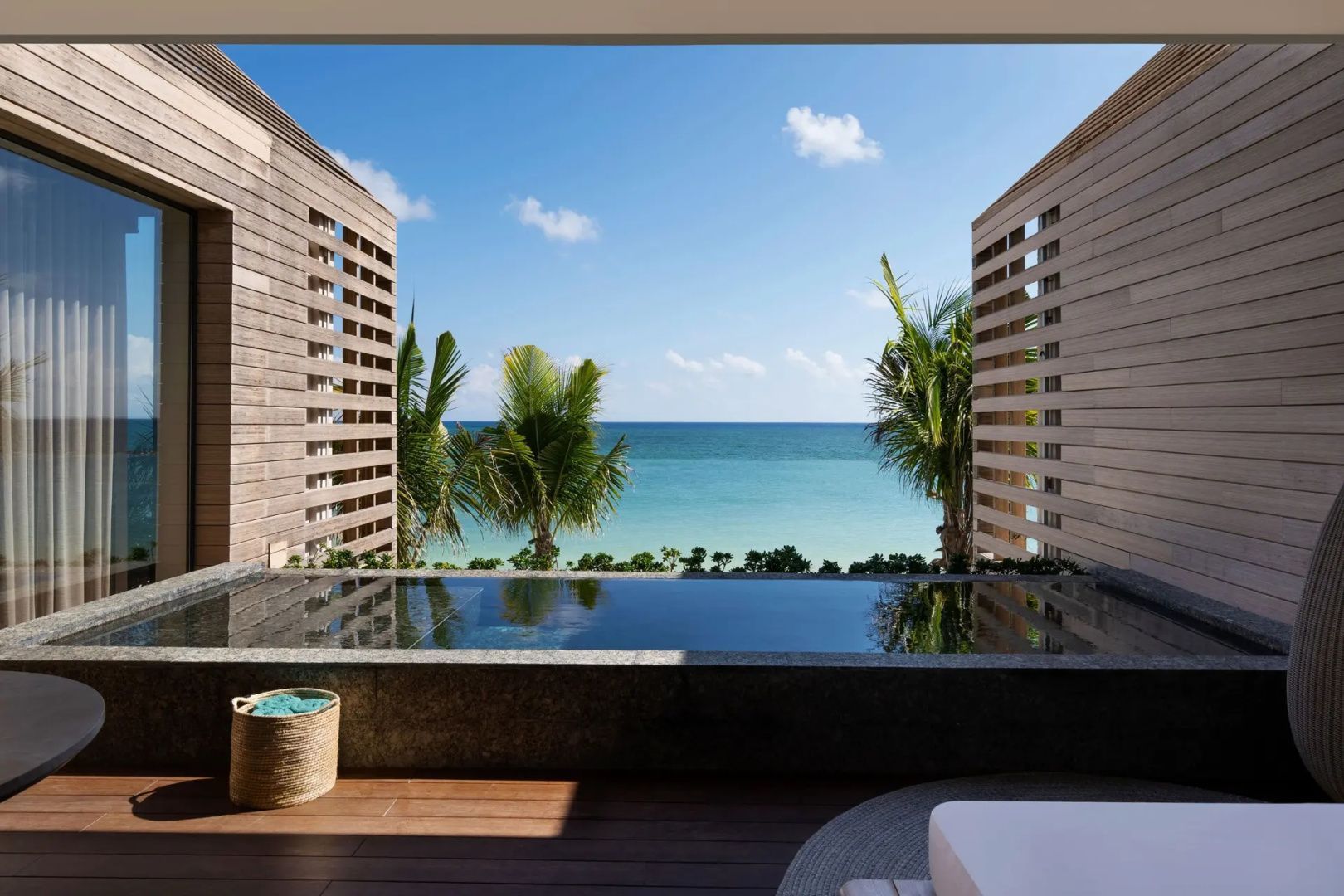 Schmaler Pool auf einer Holzterrasse mit Ausblick auf grüne Palmen und das blaue Meer.