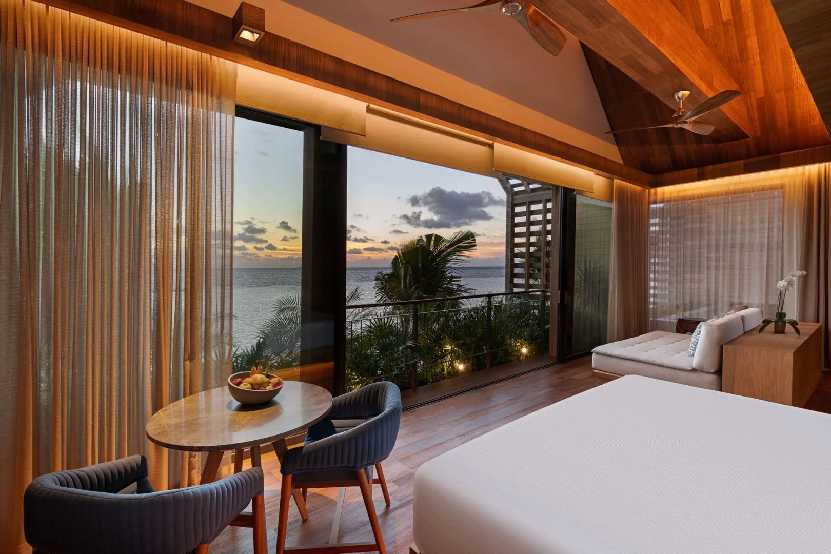 Sitzbereich im Hotelzimmer mit Blick durch große Fenster auf das Meer bei Sonnenuntergang.